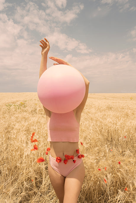 POPPIES IN THE WIND: A woman in a pink crop top and matching underwear stands in a field of golden wheat, holding a large pink balloon in front of her face. Red poppy petals are scattered around her and a few are around her waist.