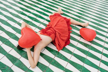 RAIN IN LOVE: A woman in a red dress lays on her stomach on a geometric tile floor. She is holding a red heart-shaped balloon over her face. Another red balloon lies beside her. The white and green tile pattern creates a visually interesting backdrop.