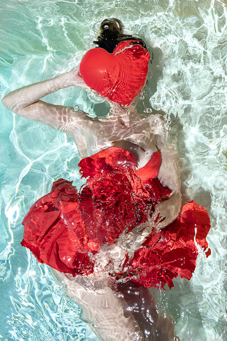 WATER IN LOVE: A woman wearing a red dress holds a red heart-shaped object underwater in a clear pool. The light shines through the water, creating a shimmering effect. The womans dark hair flows around her head.