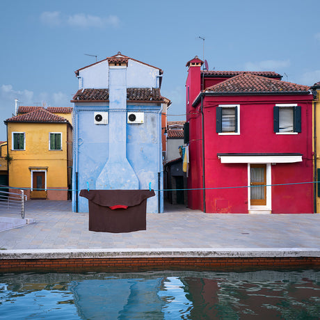 CAPTAIN: A colorful house facade in Italy, with a blue, red and yellow building. A clothesline hangs across the front of the building, with a brown t-shirt hanging on it. A canal with reflecting water can be seen in the foreground.