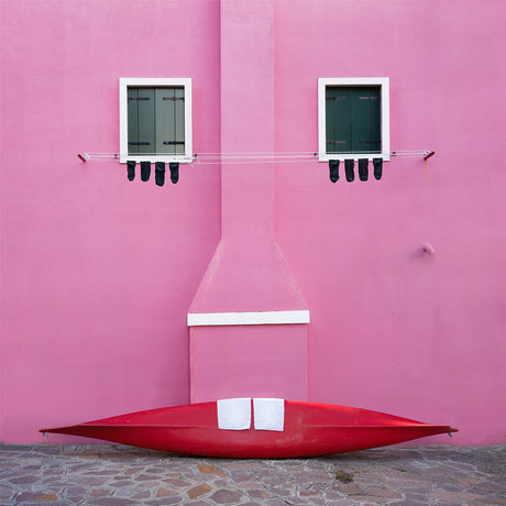 CASA ROSA: A red canoe rests against a bright pink wall in Burano, Italy. Two windows with white frames and green shutters are visible above the canoe. A clothesline with black socks hanging on it is strung between the two windows.