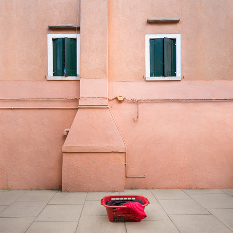 CASALINGA: A photograph of a pink wall with a laundry basket in front of it. The wall has two windows with green shutters on them. There is a pipe sticking out of the wall.