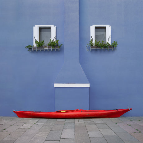 SIGNOR SORRISO: A red kayak rests against a blue wall with two windows with white trim. The windows have white shutters and are adorned with potted plants. The wall has a white trim and a chimney. The scene is simple, but evokes a sense of tranquility and relaxation.