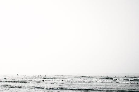 DITCH PLAINS 1: A black and white photograph of a group of people surfing in the ocean. The water is choppy and there is a light mist in the air. The sky is a bright white and the sun is not visible. The photo has a calming and minimalist feel.