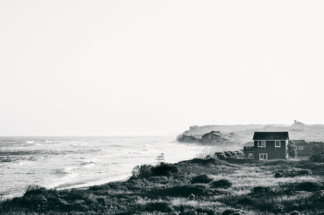 DITCH PLAINS 2: A black and white photograph of a scenic coastal view. The ocean stretches out in the distance, with waves crashing on the sandy shore. A house sits on a grassy hill overlooking the beach. The sky is clear and bright, with a few wispy clouds.