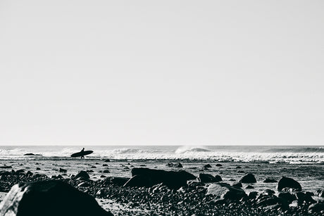 DITCH PLAINS 3: A black and white photograph captures a lone surfer walking across a rocky beach with their surfboard. The scene is backlit by the setting sun, creating a dramatic silhouette. The ocean waves are crashing in the distance, and the sand is covered in smooth, dark stones.