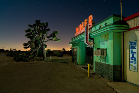 DINER: A neon sign illuminates a classic green diner in the Mojave Desert under a starry night sky. A Joshua Tree stands tall in the foreground, providing a sense of the desolate landscape. The diners bright lights offer a beacon of warmth and hospitality in the otherwise barren desert scene.