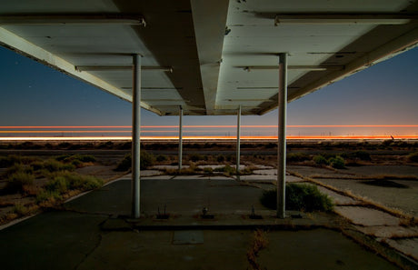 CA-58: An abandoned gas station canopy at dusk. The canopy is supported by several concrete pillars and the concrete floor is cracked and overgrown with weeds. The image captures a feeling of solitude and decay.