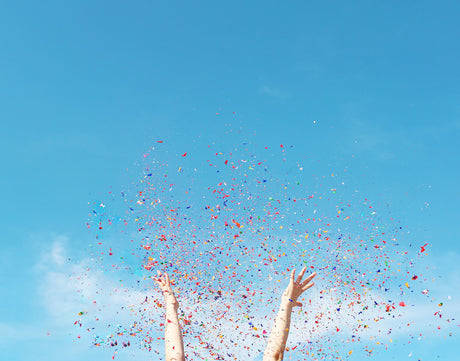 A YEAR OLDER: A person throws confetti upward into the air, celebrating and enjoying the moment. The confetti is a bright mix of colors, and the sky is a vibrant blue, signifying a joyous occasion. The image captures a sense of excitement and happiness.