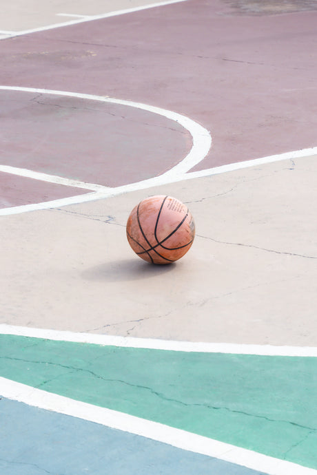 ALTURAS: A basketball rests on a colorful outdoor court, featuring a mix of pink, green, and white painted lines. The basketball is positioned near the center of the court, with no visible players or other objects in the image.