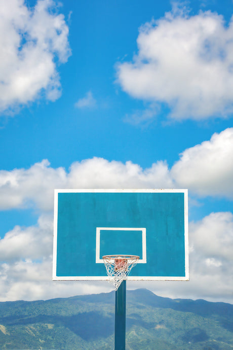 CIELO: A blue basketball hoop is set against a backdrop of a bright blue sky with fluffy white clouds. The hoop is positioned in front of a green mountain range in the distance. The image is a simple and peaceful depiction of a basketball court.