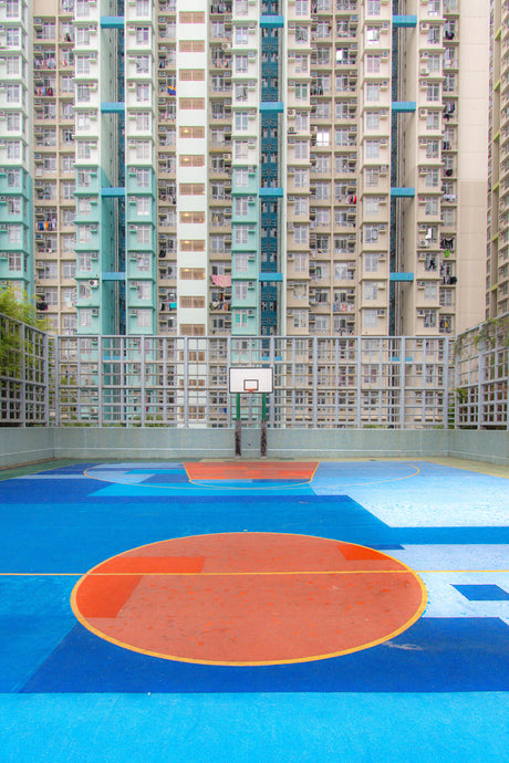 DEEP WATER: A view of an outdoor basketball court in Hong Kong, featuring a blue and orange painted court with a hoop and net. The court is situated in front of a large apartment complex, with multiple buildings visible in the background.