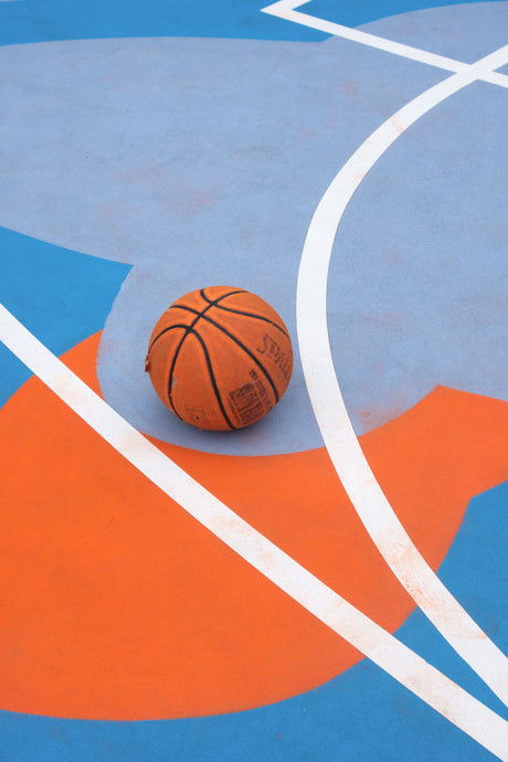 EWING: A close-up shot of a basketball resting on a colorful outdoor basketball court. The ball is positioned at the intersection of white lines, with the court painted in shades of blue and orange.