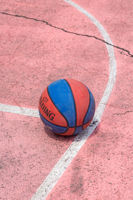 FRUIT ROLL UP: A close-up image of a basketball on a pink outdoor court with white lines. The ball is lying near the intersection of two lines and the surface of the court is slightly weathered with a few cracks.