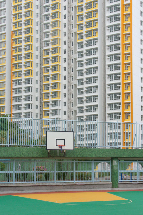 GREEN EYES 3: A basketball court with a hoop and backboard sits in front of a tall apartment building in Hong Kong. The court is surrounded by a fence and the building is visible behind it.