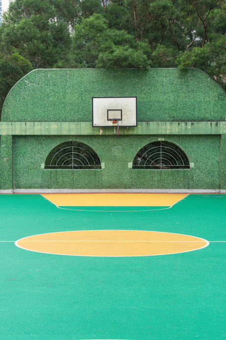 GREEN EYES 1: A green basketball court with a hoop and net in a park in Hong Kong. The court has yellow lines painted on it, and the hoop is mounted on a green wall. There are large trees surrounding the court.