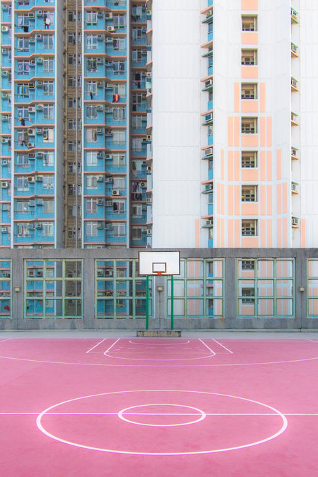 HONG KONG 194: A brightly colored basketball court is situated in front of a tall apartment building in Hong Kong. The court features a yellow, orange, white, and blue design with a basketball hoop at the far end of the court.