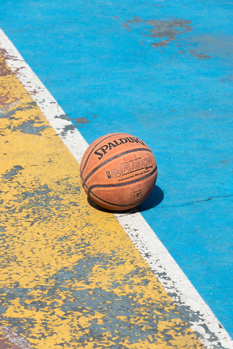 MTA: A weathered basketball sits on a cracked and faded yellow and blue outdoor basketball court. The ball is a standard size basketball with the Spalding logo visible.