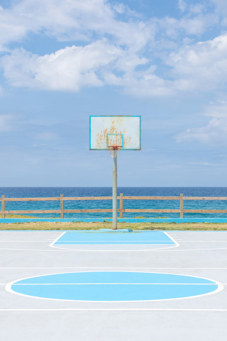 MIRAMAR: An outdoor basketball court sits beside a wooden fence overlooking a calm, blue ocean. The court is painted in white and light blue, with a basketball hoop on a pole in the center. White clouds are scattered across a bright blue sky.