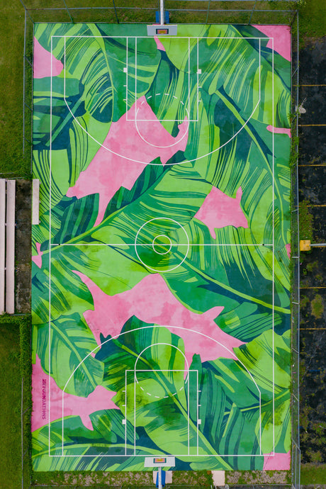 SELVA DE PUERTO RICO: This image shows an aerial view of a basketball court. The court has been painted with a tropical theme, featuring large green leaves on a pink background. The white lines of the basketball court are clearly visible, and there is a basket at each end of the court. The court is surrounded by green grass.