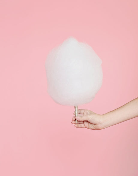 BON PUF: A single hand holds a large, fluffy cotton candy on a stick against a plain pink background. The cotton candy is white and round, taking up most of the space in the image. The hand is lightly holding the stick, and the background is a soft pink.