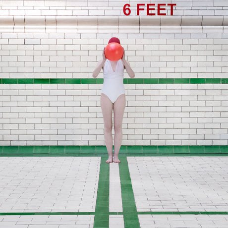 SIX FEET: A woman is standing in a tiled pool area, wearing a white swimsuit and a red bathing cap. She is holding a red ball in front of her face. The sign above her reads 6 FEET. The tiled walls are white with green trim, and the floor has green and white tiles.