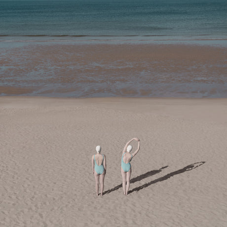 SUNDAYS AT THE BEACH: This image features two women wearing swimsuits and swim caps, standing on a sandy beach with their backs to the camera. They are stretching their arms overhead, likely getting ready for a swim in the ocean. The beach appears to be deserted, with only the two women and their shadows visible in the photo. The ocean is in the background, and the sky is a muted blue.