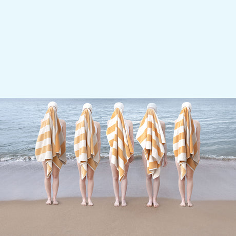 SWIM TEAM AT TYNINGHAME: Five people are standing on a sandy beach, facing away from the camera, with their bodies obscured by yellow and white striped towels. The beach is empty, and the ocean is calm in the background. The scene is minimalist and somewhat mysterious.