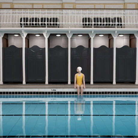 SWIMMER IN YELLOW: A woman in a yellow swimsuit and white swim cap sits on the edge of an indoor swimming pool. The pool is surrounded by black changing rooms and a white railing. There is a sign on the edge of the pool that reads 1.2m.