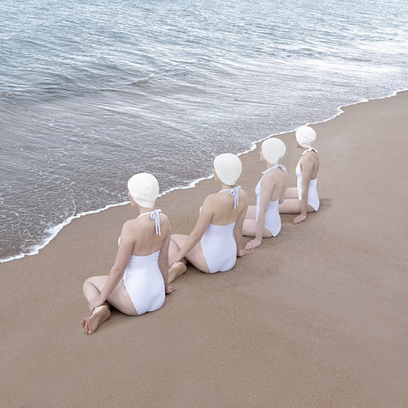 TYNINGHAME: Four women are sitting on a sandy beach, facing the ocean, wearing white swimsuits and white swim caps. They are all sitting in a row, facing forward. The water is calm and the sky is clear.