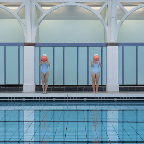 WATER POLO AT WARRENDER: This image depicts two women standing at the edge of a swimming pool. They are both wearing blue swimsuits and holding red balls in front of their faces. The pool is surrounded by white pillars and blue walls. The setting is likely an indoor pool.