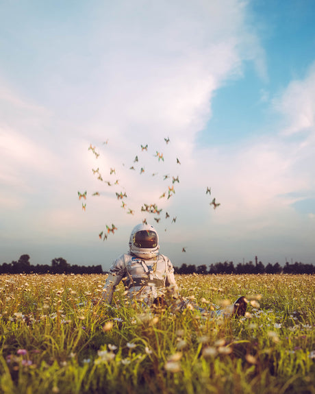 ASTRO BUTTERFLIES: An astronaut, wearing a white spacesuit, sits in a field of wildflowers under a blue sky with white clouds. Butterflies fly around the astronauts head, creating a whimsical scene that contrasts the astronauts serious demeanor and the vastness of space. The astronauts presence in the field creates a sense of wonder and possibility, as if he has come from another world.