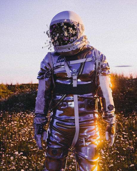 ASTRO FLOWER MASK: A full-body shot of an astronaut standing in a field of white wildflowers at sunset. The astronaut is wearing a silver spacesuit and a helmet filled with flowers. The sun is setting behind the astronaut, creating a warm, golden glow.