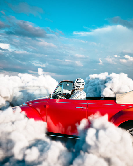 ASTRO FLY 1: A red convertible car drives through a field of fluffy white clouds on a sunny day. The driver, wearing a silver helmet, is behind the wheel, enjoying the ride. The car is moving fast and leaving a trail of smoke behind it. The clouds are a beautiful contrast to the bright blue sky.