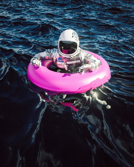 ASTRO LOST AT SEA: An astronaut in a white helmet and silver spacesuit floats on a pink inflatable ring in a body of blue water. The astronaut appears to be enjoying a relaxing float in the water, with the sun reflecting on the surface.