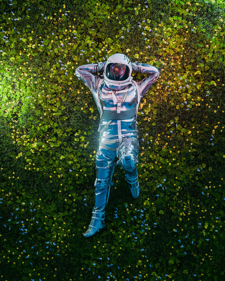 ASTRO NEW YEARS: This image shows an astronaut in a silver spacesuit lying on their back in a field of green grass and flowers. The astronaut is wearing a white helmet with a visor and has their hands behind their head. The background is a field of flowers with a soft, blue-toned glow. This creates a surreal and dreamy scene.