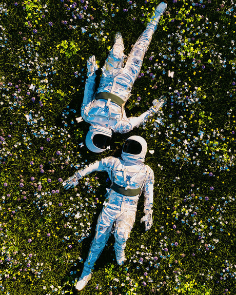 SATURDAY AFTERNOON: This image shows two astronauts lying in a field of wildflowers, enjoying a break from their space travels. They are wearing white spacesuits and helmets, and their arms and legs are relaxed. The field is full of vibrant green grass and colorful flowers, creating a peaceful and serene atmosphere.