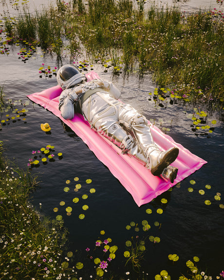 THE POND 1: An astronaut in a silver spacesuit lays on a pink inflatable float in a body of water. The water is surrounded by green grass and white and pink flowers. The astronaut appears to be taking a break from space travel.