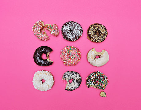 CHOCOLATE FOR THE WIN!: Nine donuts, each with a different glaze, are arranged in a grid pattern on a pink background. Some of the donuts have bites taken out of them, adding a playful touch to the image. The variety of colors and textures of the donuts creates a visually appealing composition.