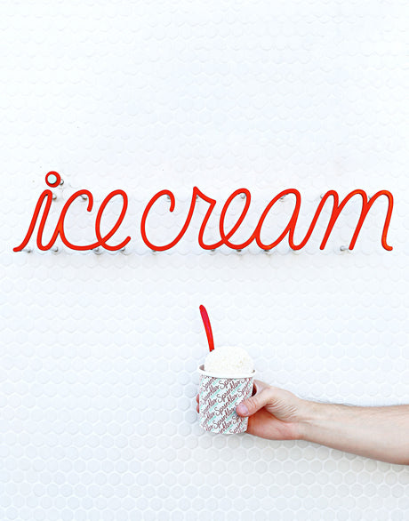 CRÈME DE LA CRÈME: A hand holds a cup of ice cream with a red spoon in front of a white wall with a red neon sign that says ice cream.