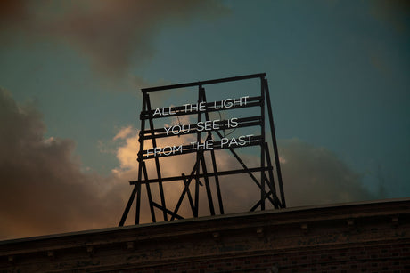 ALL THE LIGHT YOU SEE: A photograph capturing a weathered wooden frame sign with a neon message reading All The Light You See Is From The Past against a cloudy sky. The sign is situated atop a brick building, emphasizing the idea that our perceptions are shaped by the past.