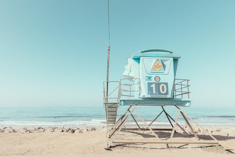 10 STATE PARKS: A blue lifeguard tower stands on a sandy beach, overlooking the calm ocean. The tower is marked with the number 10, indicating its position along the coastline. The clear, blue sky and tranquil water create a serene and picturesque setting.