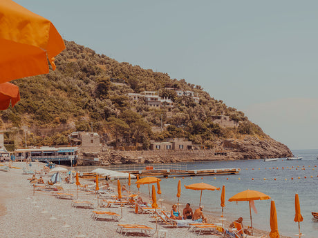 AMALFI COAST: A picturesque scene of a beach in Italy, featuring bright orange umbrellas lining the shore. The beach is filled with people relaxing under the sun, enjoying the beautiful day. The calm waters of the ocean and the lush green hillside behind the beach create a serene and idyllic backdrop.