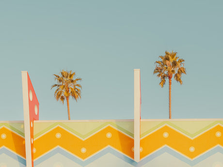 AMUSEMENT PARK SANTA CRUZ: A photograph of a colorful wall with two palm trees in front of it. The wall features a chevron pattern in yellow, green, blue, and white. The sky is a bright blue, suggesting a sunny day. The palm trees are in focus, and the wall is slightly blurred.