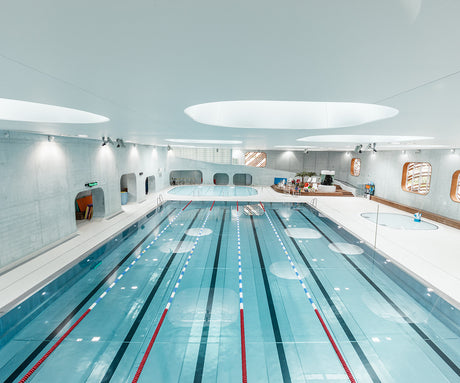 AQUAZENA: A photograph of an indoor swimming pool, showcasing a long, laned pool with red lane dividers, a circular pool, and a white tiled floor. The pool is lit by overhead lights, and the ceiling features unique oval-shaped skylights.