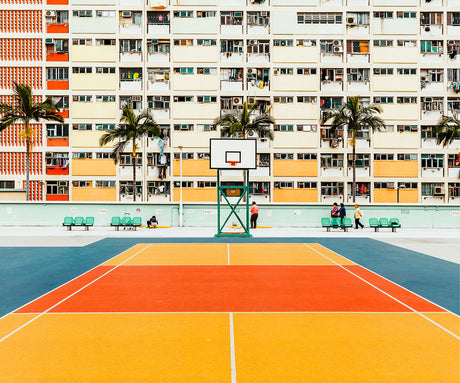 BASKETBALL HONG KONG: A view of a brightly colored basketball court in front of an apartment building in Hong Kong. The court features a blue, orange, and yellow color scheme, and the building is adorned with a mix of yellow and orange. The image features a few people walking and sitting on the benches in the background.