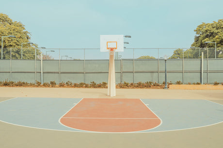 BEVERLY HILLS COURT: A view of an empty basketball court on a sunny day. The court has a blue and orange colored key, and a white backboard with a net hangs from a white pole in the center of the court. A chain-link fence surrounds the court.