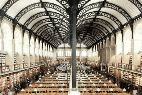 BIBLIOTHEQUE SAINTE GENEVIEVE: A photo of the interior of a large library reading room with rows of wooden tables and chairs. The room is tall, with ornate arches and a black metal ceiling. Sunlight streams in through windows along the side walls.