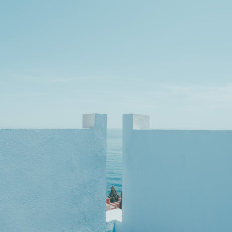 BLUE WALL: A photo of a white wall with a gap that reveals a view of the blue ocean in the distance. The image is taken from a high vantage point, likely from a rooftop or balcony, showcasing the architectural elements of a coastal town. The bright blue sky and the minimal composition create a sense of serenity and tranquility.