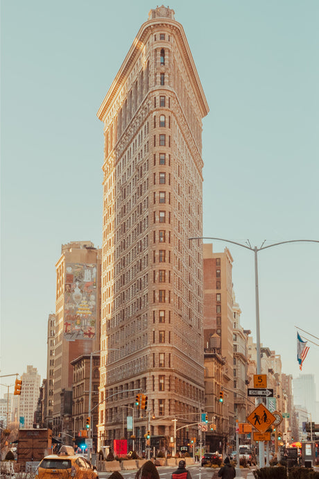 FLATIRON: A classic view of the Flatiron Building in New York City, showcasing its distinctive triangular shape and towering presence. The building stands tall against a clear blue sky, illuminated by the warm glow of the sun. The scene is bustling with activity, with pedestrians crossing the street and cars navigating the citys traffic. The Flatiron Building, a symbol of the citys architectural heritage, is a popular tourist destination and a must-see for anyone visiting New York City.